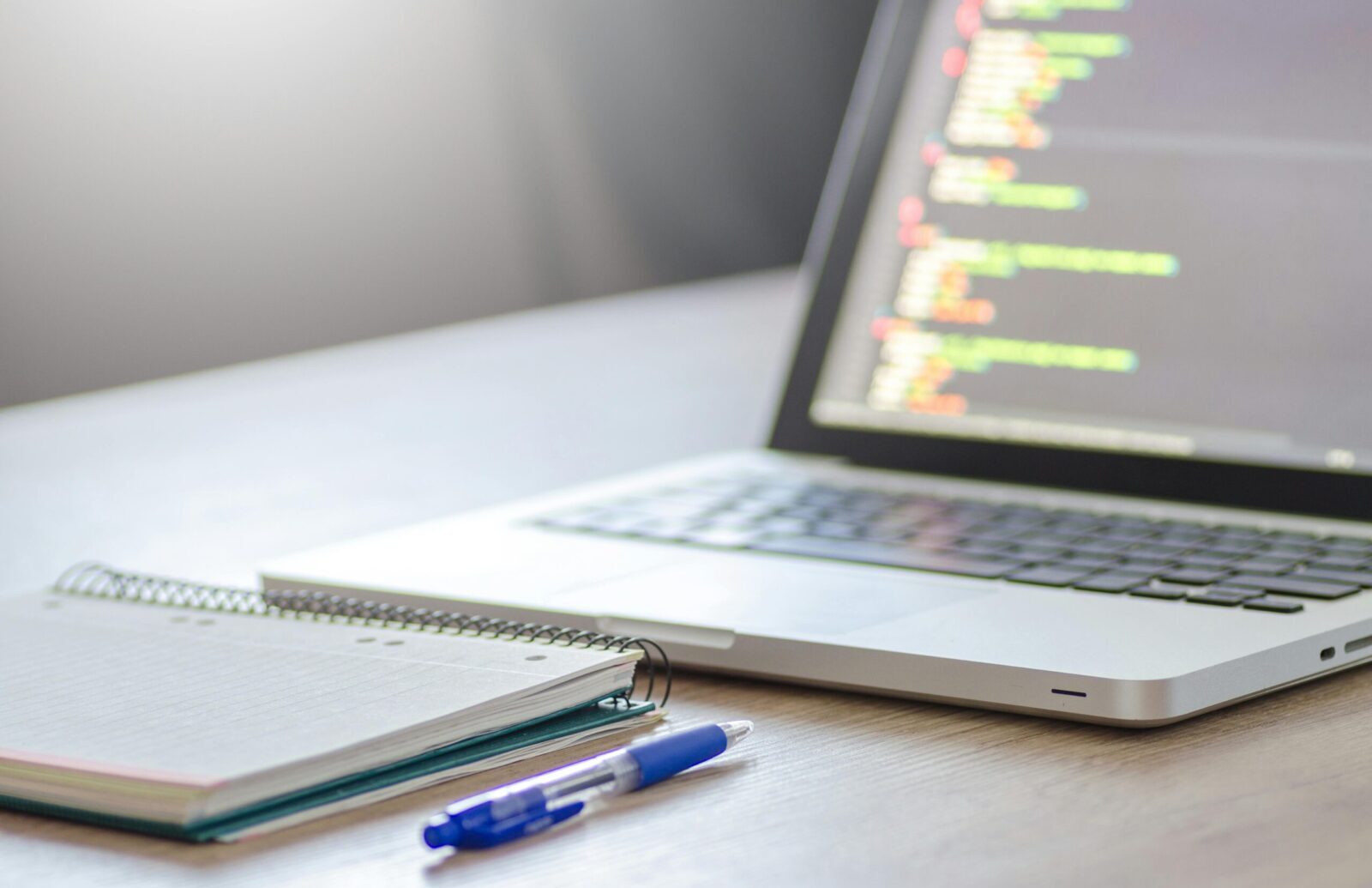 Open laptop with programming code on screen next to a notebook and pen on a desk.